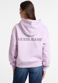 Felpa cappuccio lavanda con vestibilità comoda, presenta il logo "GUESS JEANS" in bold nero, polsini e cintura a coste. Abbinata a jeans blu.