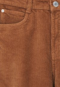 Kordtyg i en rik brun färg med vertikala ribbor, en framficka och en metallknappdetalj. Mjuk textur.