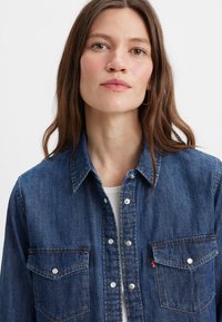 Chemise en denim bleu foncé avec un col, deux poches poitrine et fermeture à boutons. Présente des coutures légères et une étiquette logo Levi's.