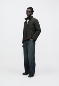 Schwarze gesteppte Jacke mit Stehkragen, ausgestattet mit einem Frontreißverschluss und Seitentaschen. Kombiniert mit dunklen, weit geschnittenen Jeans und schwarzen Schuhen.