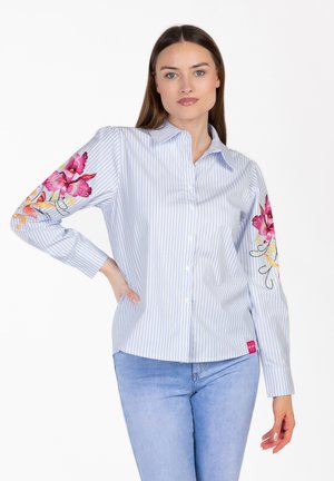 Mujer vistiendo una camisa de botones a rayas azul claro con bordado floral rosa en las mangas y jeans azul claro, de pie con una mano en la cadera.