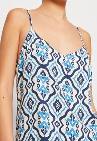 Žena v bezramenném topu s tenkými popruhy a modrým geometrickým vzorem na bílém pozadí.