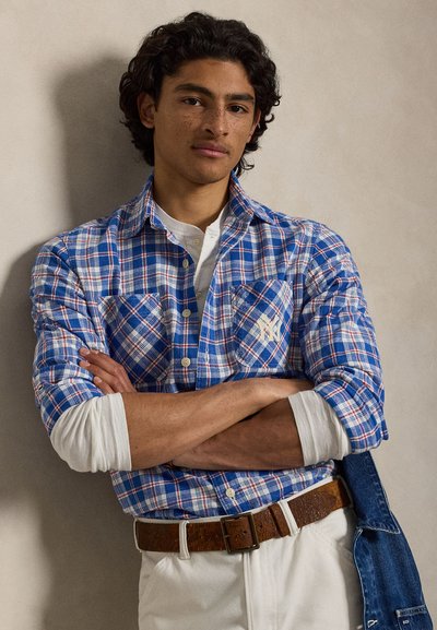 Mladić s kovrčavom kosom, nosi plavu kariranu košulju preko bijele majice dugih rukava, bež hlače i smeđi remen, stoji s prekriženim rukama.