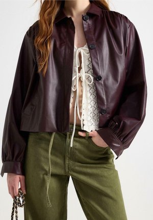 Femme portant une veste en cuir marron foncé boutonnée sur un top blanc à lacets, associée à un pantalon vert olive, tenant un sac à main orné de chaînes.