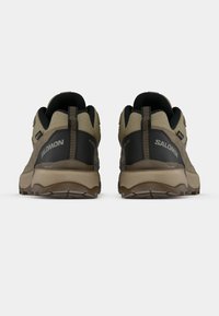 Καφέ και μαύρα παπούτσια πεζοπορίας με υφασμένο πάνω μέρος, ενισχυμένη πτέρνα, καμπυλωτή σόλα και εμφανές λογότυπο στο πίσω μέρος της πτέρνας.