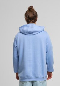 Felpa azzurra chiara realizzata in tessuto morbido, con un ampio cappuccio, maniche lunghe e una vestibilità rilassata con dettagli di cucitura visibili.