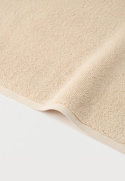 Μαλακή πετσέτα σε απαλό μπεζ χρώμα με υφή από επαναλαμβανόμενο ελαστικό ύφασμα, μερικώς διπλωμένη σε λευκό φόντο.