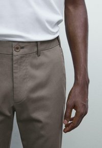 Man in een lichtbruine broek en een wit shirt, met de rechterarm en hand ontspannen langs het lichaam tegen een effen achtergrond.