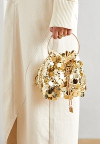 Zlatá kabelka typu bucket s flitry, kulatým kovovým madlem, stahovacím zavíráním a řetízkovými doplňky. Struktur surface s kruhovými ozdobami.