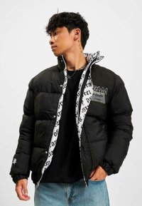 Jachetă puffer neagră cu fermoar frontal, manșete elastice și căptușeală interioară cu modele de text alb. Include o etichetă cu logo pe piept.
