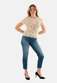 Top de encaje beige con patrones intrincados, combinado con jeans skinny azul claro y tacones altos negros. El top tiene un escote redondo y mangas cortas.