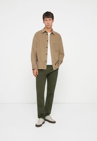 Light brown plaid button-up shirt over a white t-shirt, paired with green pants and cream sneakers, standing against a white background.