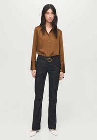 Woman standing wearing a brown satin blouse, dark jeans with a brown belt, and white pointed-toe shoes against a plain background.