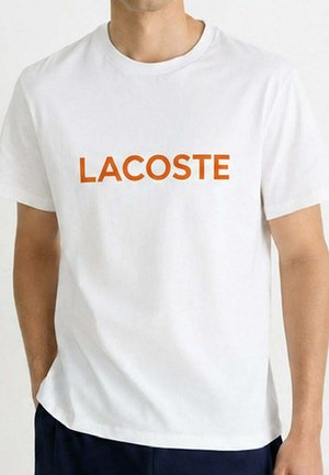 Hombre con camiseta blanca con texto naranja "LACOSTE" en el pecho y pantalones oscuros, de pie con las manos en los bolsillos.