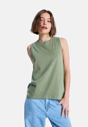 Młoda kobieta z krótkimi brązowymi włosami, ubrana w oliwkowozielony, bezrękawnik i jasnoniebieskie dżinsy, pozująca na białym tle.