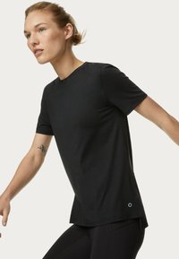 Camiseta negra de manga corta con cuello redondo, tejido suave, corte relajado y aberturas laterales, con un logo circular cerca del dobladillo.
