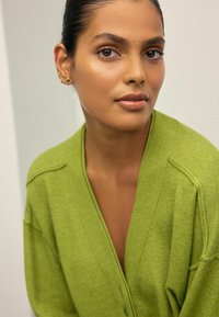 Grüne Strickjacke aus strukturiertem Strickstoff mit tiefem V-Ausschnitt, vorderen Knöpfen und überschnittenen Schultern. Nahaufnahme einer Person, die sie trägt.
