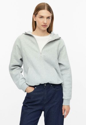 Jeune femme portant un sweat à capuche zippé gris clair par-dessus un t-shirt blanc et un jean bleu foncé, debout avec une main dans la poche sur fond blanc.