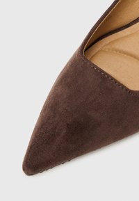 Scarpa in suede marrone con punta a freccia e design con tagli. La soletta è beige e le cuciture lungo il bordo sono visibili, aumentando la texture.