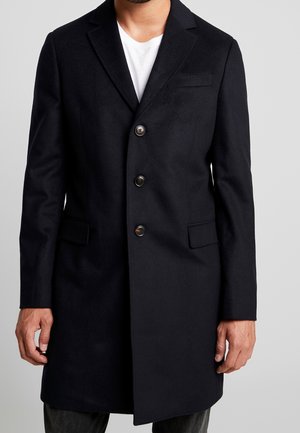 Mand iført en sort tre-knaps frakke over en hvid skjorte, stående foran en ensartet lys grå baggrund.