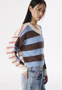 Pull-over tricoté avec un col en V, présentant de larges rayures horizontales marron, bleu et crème, avec des accents rouges sur une manche. Tissu texturé.