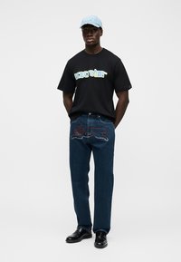 Μαύρο T-shirt κοντομάνικο με την εκτύπωση "icecream" σε κίτρινο και μπλε χρώμα· σκούρο μπλε τζιν με κόκκινες και ασημί διακοσμήσεις· ανοιχτό μπλε καπέλο.