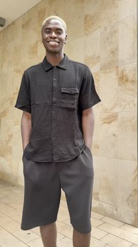 Camisa preta de manga curta com fecho de botão e um único bolso, combinada com calções escuros até ao joelho; tecido suave e ajuste descontraído.