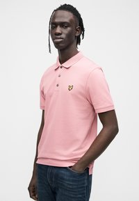 Roosa polo-särk pehme tekstuuriga, millel on kolme nupu plastron ja kollane tikitud logo. Kandmisega tumedate siniste teksadega.