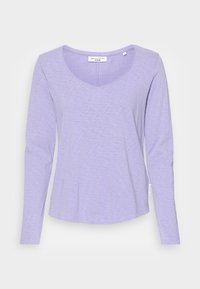 Długo rękawowy top w jasnym lawendowym kolorze, z dekoltem w kształcie litery V i miękkim, teksturowanym materiałem. Dobrany brzeg i metka z logo po boku.