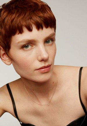 Collar de cadena de oro con un pequeño colgante, llevado sobre piel desnuda; el sujeto tiene el cabello corto, de color castaño rojizo, y viste una blusa negra.