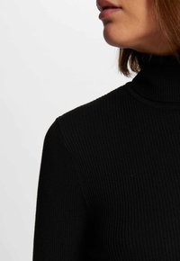 Zwarte geribbelde coltrui met een aansluitend ontwerp en textuurstof, met een hoge kraag en lange mouwen. Achtergrond is vlak wit.