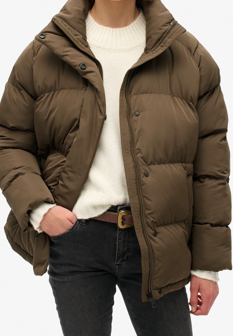 Ruskea toppatakki, jossa korkea kaulus, edessä vetoketju ja napeilla kiinnitettävä läppä. Päällä luonnonvalkoinen neule, yhdistetty tummiin farkkuihin ja ruskeaan vyöhön.