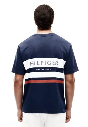 Férfi visel tengerészkék rövid ujjú inget, fehér, fekete és piros vízszintes csíkokkal és „HILFIGER MARINA CLUB” felirattal a háton.