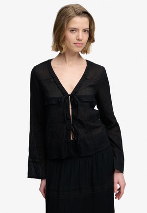Femme aux cheveux blonds courts portant un chemisier noir transparent noué sur le devant et une jupe assortie, debout devant un fond blanc.