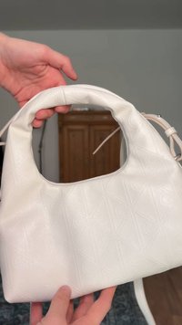 Λευκή δερμάτινη τσάντα χειρός με ανάγλυφα γεωμετρικά σχέδια, κρατημένη με δύο χέρια, που δείχνει τη κυρτή πάνω λαβή και το ρυθμιζόμενο λουρί της.