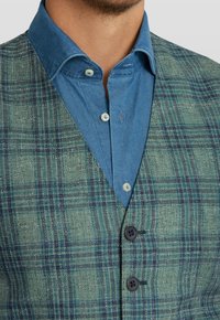 Grön rutig väst med en strukturerad väv, med två mörka knappar. Bärs över en ljusblå skjorta med knappar.