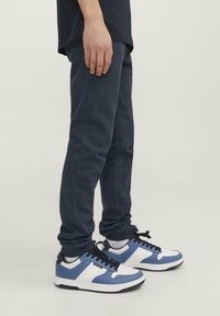 Pantalones de chándal azules fabricados en un tejido suave, combinados con zapatillas blancas y azules con cordones negros y una suela texturizada. Vista en ángulo que muestra el ajuste.