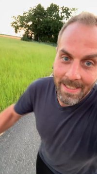 Mann mit Bart und blauen Augen, der ein dunkles Hemd trägt und ein Selfie macht, während er auf einem gepflasterten Weg neben grünem Gras und Bäumen spaziert.