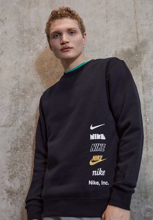 Felpa nera con vari loghi Nike in bianco e oro, scollo rotondo e polsini a costine. Sfondo grigio con texture in cemento.
