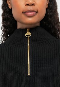 Sweater preto de malha com gola alta e detalhe em zíper dourado no pescoço. A textura é canelada, proporcionando uma aparência estruturada.