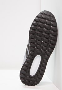 Grå atletisk skosula med rundad svart gummitread, texturerad yta och en vit logotyp mot en ljus bakgrund. Sidodekor i grått.