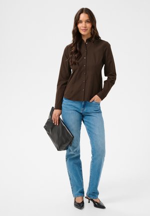Femme aux longs cheveux bruns portant une chemise boutonnée marron foncé, un jean bleu, des talons noirs et tenant une grande pochette noire.