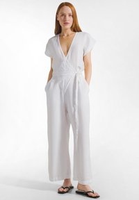 Kvinna med rött hår iförd en vit linnesuit i omlottstil med fickor och svarta flip-flop-sandaler stående mot en vit bakgrund.