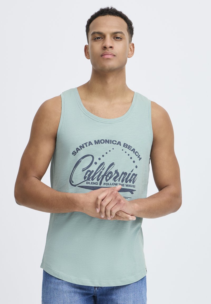 Lys grønn tanktopp med rund hals og grafisk trykk som sier "Santa Monica Beach California." Kombinert med blå jeans.