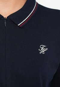 Tengerészkék galléros póló texturált anyagból, cipzáras gallérral, valamint piros-fehér csíkos díszítéssel a galléron. Fehér hímzett logóval.