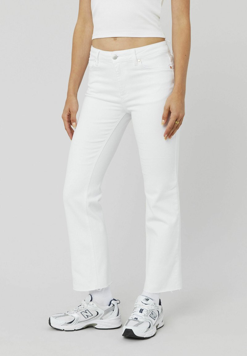 Jeans évasés blancs en denim, avec une taille mi-haute, des poches avant et un léger effilochage à l'ourlet. Assortis à des baskets blanches.