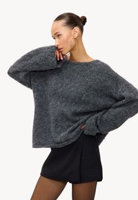 Pull en tricot gris, surdimensionné, fabriqué à partir d'un matériau doux, avec un col rond et des épaules tombantes, associé à une jupe noire.