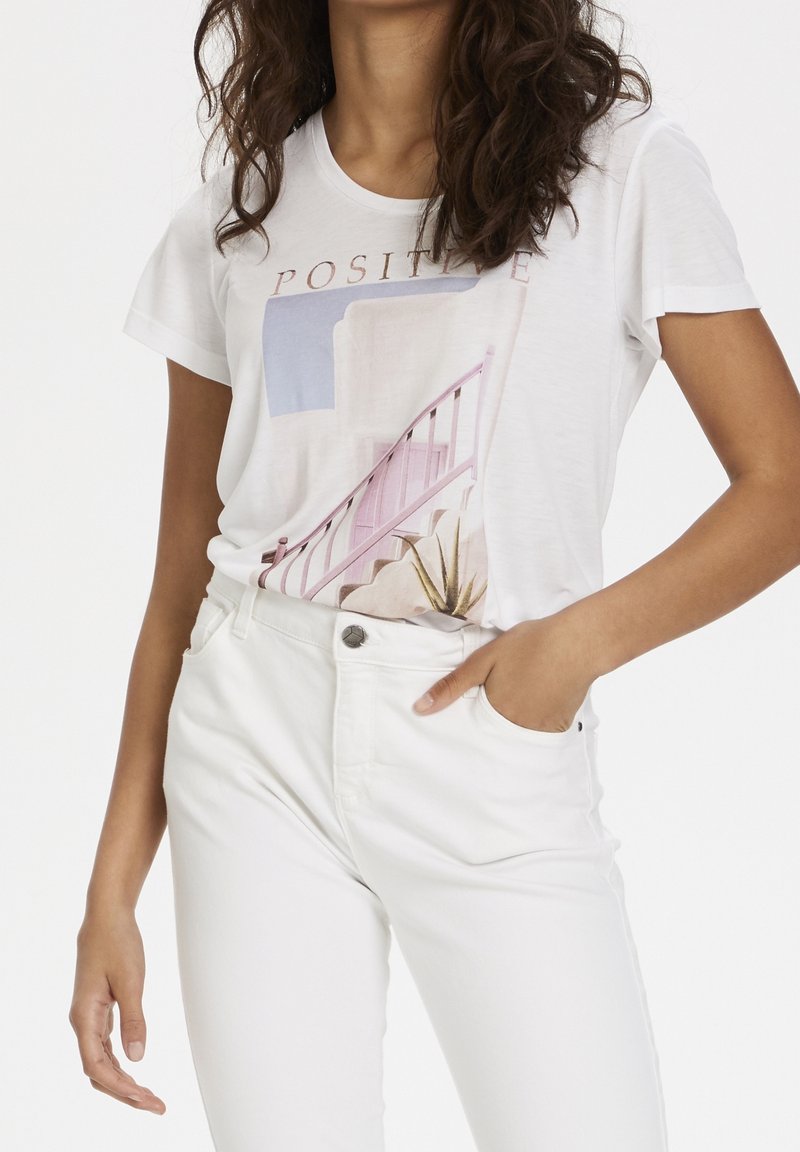 Mujer vestida con pantalones blancos y una camiseta gráfica blanca con la palabra "Positivo" y un diseño de escalera en tonos pastel.