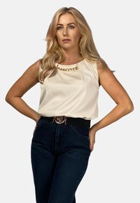 Kremowa bluzka bez rękawów z łańcuszkiem przy dekolcie, zestawiona z męskimi ciemnoniebieskimi dżinsami z wysokim stanem oraz czarnym paskiem ze złotą klamrą.