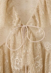 Gros plan d'un tissu en dentelle florale beige avec un nœud noué au centre, fait de fins cordons beiges.